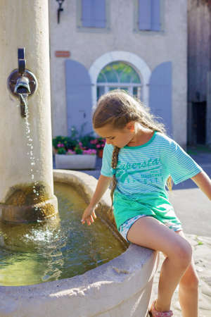 Girl looks at the water of old fountain on the typical square of small European villages with cozy streets and stone houses, summer vacations, family travel, beautiful summertimeの写真素材