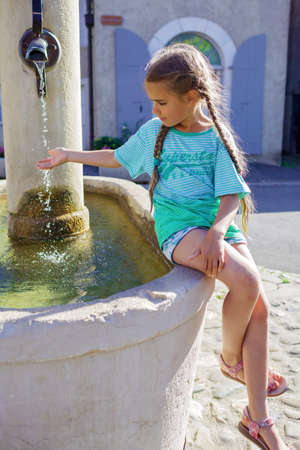 Girl puts her hand in the water of old fountain on the typical square of small European villages with cozy streets and stone houses, summer vacations, family travel, beautiful summertimeの写真素材
