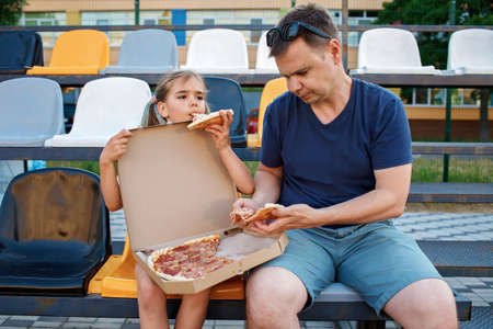 Father with daughter, soccer fans, watching football match, eating pizza and cheering for local team at stadium, real emotions, sports event and fan supporting, outdoor lifestyle, street foodの写真素材