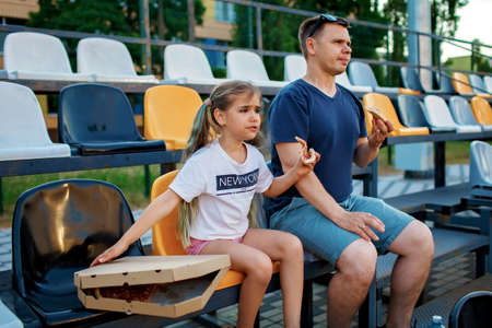 Father with daughter, soccer fans, watching football match, eating pizza and cheering for local team at stadium, real emotions, sports event and fan supporting, outdoor lifestyle, street foodの写真素材