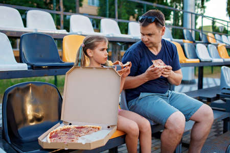 Father with daughter, soccer fans, watching football match, eating pizza and cheering for local team at stadium, real emotions, sports event and fan supporting, outdoor lifestyle, street foodの写真素材