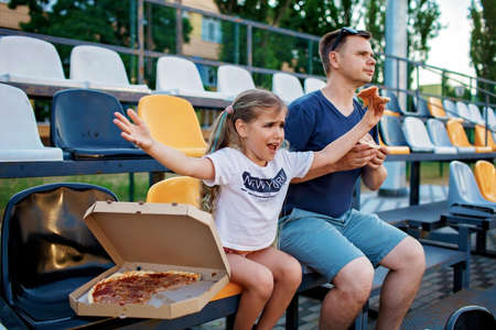 Father with daughter, soccer fans, watching football match, eating pizza and cheering for local team at stadium, real emotions, sports event and fan supporting, outdoor lifestyle, street foodの写真素材