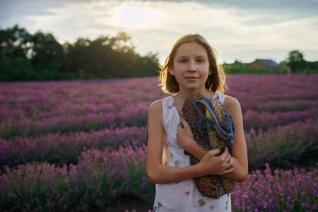 Teenage girl holds cotton reusable net bag with a bottle of water and French baguette in lavender field. Teenager walks and ready for picnic in purple flowers at sunset. Summertime, travel, vacationの写真素材