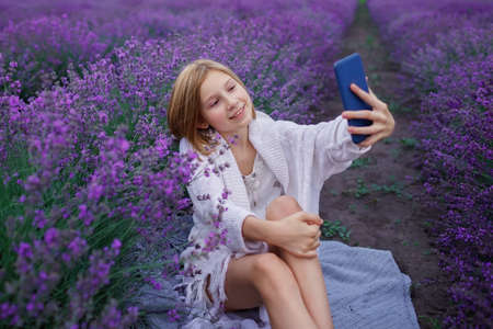 Teenage girl enjoys lavender field and make photo with smartphone. Gen Z teenager with gadget walks among purple flowers at sunset. Technology and beauty of nature, summer travel and vacationの写真素材