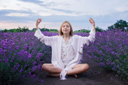 Teenage girl sits in lotus pose and enjoys scenery of lavender field. Gen Z teenager does yoga among purple flowers on beautiful sunset. Calm landscape, healthy lifestyle, summer travel and vacationの写真素材