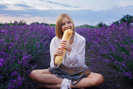 Teenage girl bites fresh French baguette in lavender field. Teenager has a picnic in purple flowers at sunset and enjoys it. Summertime, travel, vacationの写真素材