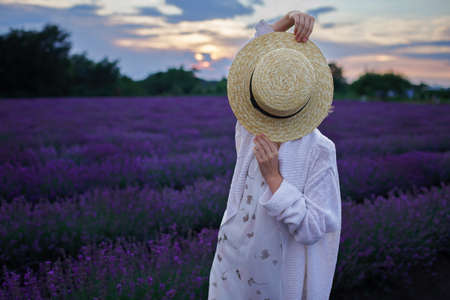 Teenage girl closes her face with straw hat in lavender field. Dreamy teenager among purple flowers at beautiful sunset. Calm landscape, faceless, escape to beauty of nature, summer travel, vacationの写真素材