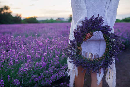 Young girl holds wreath of lavender flowers behind her back and enjoys beautiful sunset over the field. Happy teenager among purple flowers. Calm landscape, beauty of nature, summer travel, vacationの写真素材