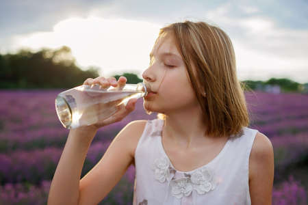 Teenage girl drinks pure water from the glass bottle at lavender field. Gen Z teenager walks in purple flowers at sunset. Water balance in heat wave. Summer weather, nature. Travel and vacationの写真素材