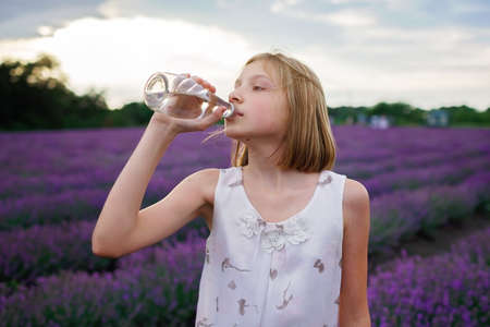 Teenage girl drinks pure water from the glass bottle at lavender field. Gen Z teenager walks in purple flowers at sunset. Water balance in heat wave. Summer weather, nature. Travel and vacationの写真素材