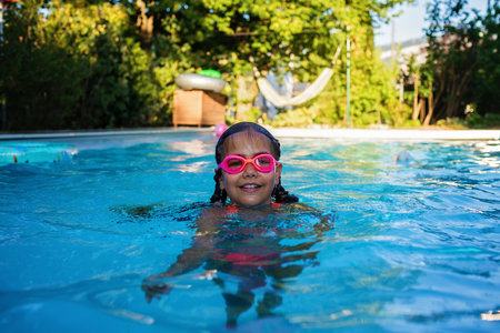 Beautiful girl with two braids in swimming suit and goggles smiles and enjoy training swim at pool in summer camp. Refreshing at heat weather, active vacation and healthy lifestyle, happy summertimeの写真素材
