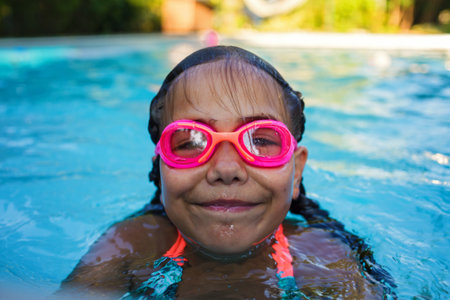 Portrait of girl with two braids in swimming suit and goggles smiles and enjoy training swim at pool in summer camp. Refreshing at heat weather, active vacation and healthy lifestyle, happy summertimeの写真素材