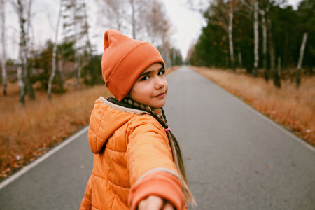 Happy girl in orange coat and hat stretches out the hand to invite and share with her the pleasure of walking through the fall forest. Dry grass and golden trees. Outdoor lifestyle. Hello autumnの写真素材