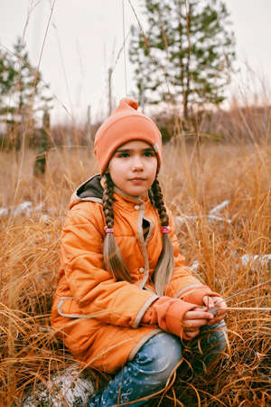 Thoughtful ten years girl in orange coat sits in fall forest among high dry grass and golden trees. Autumn vibes, outdoor lifestyle. Hello autumn, thinking and mental health conceptの写真素材