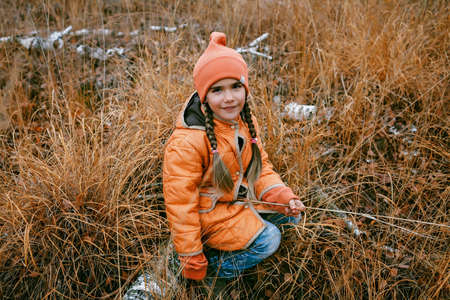 Happy ten years girl in orange coat sits in fall forest among high dry grass and golden trees. Autumn vibes, outdoor lifestyle. Hello autumn, thinking and mental health conceptの写真素材