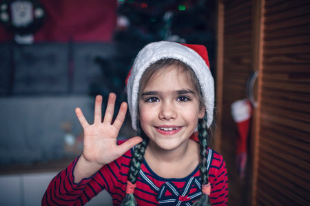 Close-up portrait of girl in Santa red hat, as if we saw her on a laptop or phone screen celebrating virtually via internet. Video call. Refugees, immigrants, distant holidayの写真素材