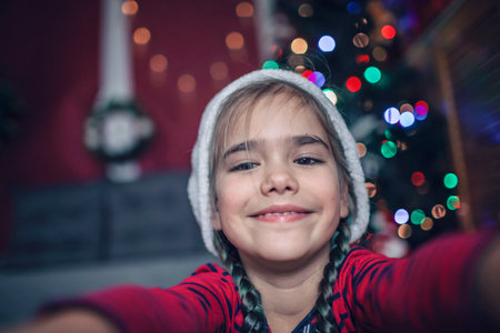 Close-up portrait of girl in Santa red hat, as if we saw her on a laptop or phone screen celebrating virtually via internet. Video call. Refugees, immigrants, distant holidayの写真素材
