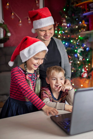 Safe online Christmas celebration. Happy family in Santa red hats celebrating with friends virtually via internet and notebook. Video call. Stay home, distant holiday, lifestyleの写真素材