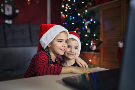Safe online Christmas celebration. Happy kids in Santa red hats with burning sparkler celebrating with friends virtually via internet and notebook. Video call. Stay home, distant holiday lifestyleの写真素材
