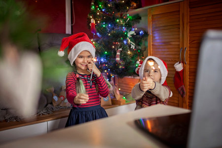 Safe online Christmas celebration. Happy kids in Santa red hats with burning sparkler celebrating with friends virtually via internet and notebook. Video call. Stay home, distant holiday lifestyleの写真素材