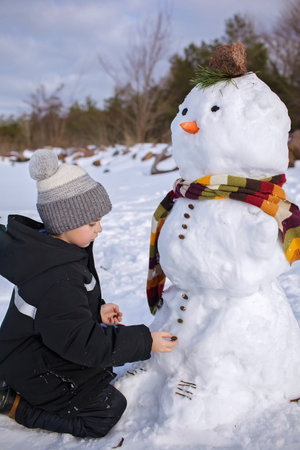 Seven years boy in knitted winter hat makes a snowman on the frozen bank of lake. Seasonal family weekend, active authentic lifestyleの写真素材