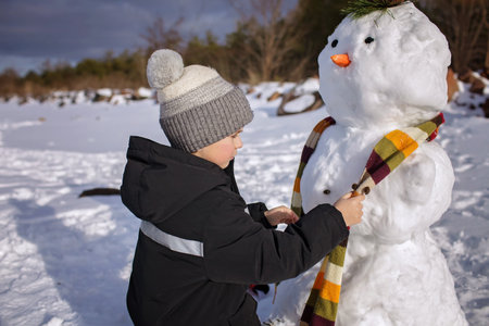 Seven years boy in knitted winter hat makes a snowman on the frozen bank of lake. Seasonal family weekend, active authentic lifestyleの写真素材