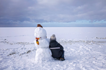 Seven years boy in knitted winter hat sits alone with snowman and looks forward. Loneliness and a friend made of snow. Winter mood on a cloudy dayの写真素材