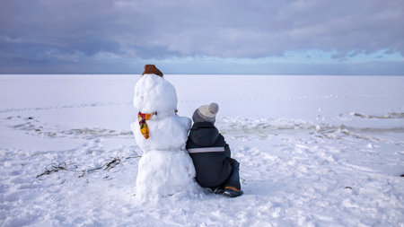 Seven years boy in knitted winter hat sits alone with snowman and looks forward. Loneliness and a friend made of snow. Winter mood on a cloudy day. Banner formatの写真素材