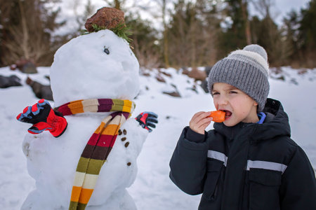 Seven years boy in knitted winter hat bites a carrot, which was the nose of snowman, smiling. Seasonal family weekend, active authentic lifestyleの写真素材