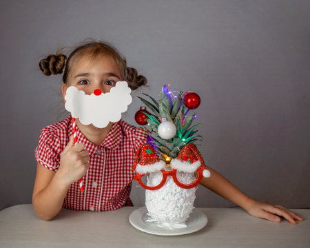 Happy cute little girl near the Santa Claus face made with white painted pineapple in festive sunglasses on the table, trendy exotic Christmas holidays over white backgroundの写真素材