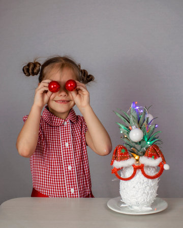 Happy cute little girl near the Santa Claus face made with white painted pineapple in festive sunglasses on the table, trendy exotic Christmas holidays over white backgroundの写真素材