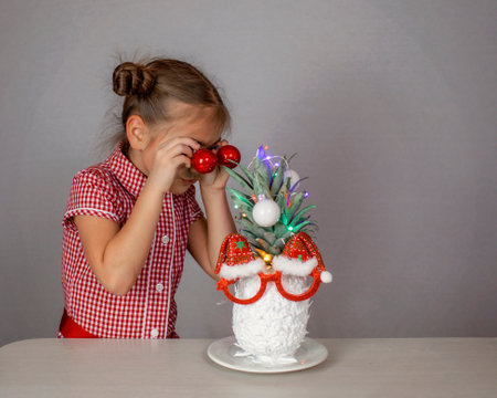 Happy cute little girl near the Santa Claus face made with white painted pineapple in festive sunglasses on the table, trendy exotic Christmas holidays over white backgroundの写真素材