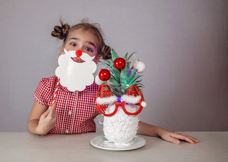 Happy cute little girl near the Santa Claus face made with white painted pineapple in festive sunglasses on the table, trendy exotic Christmas holidays over white backgroundの写真素材