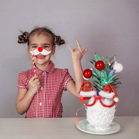 Happy cute little girl near the Santa Claus face made with white painted pineapple in festive sunglasses on the table, trendy exotic Christmas holidays over white backgroundの写真素材