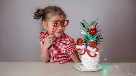 Happy cute little girl near the Santa Claus face made with white painted pineapple in festive sunglasses on the table, trendy exotic Christmas holidays over white backgroundの写真素材