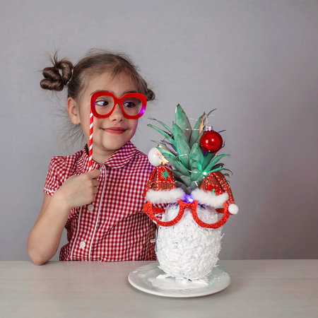 Happy cute little girl near the Santa Claus face made with white painted pineapple in festive sunglasses on the table, trendy exotic Christmas holidays over white backgroundの写真素材