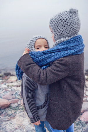 Little pretty girl in knitted grey hat hugging her frozen smaller brother during snowfall in winter, one scarf for two to keep warm in white cold weather, winter outside lifestyle close-up portraitの写真素材