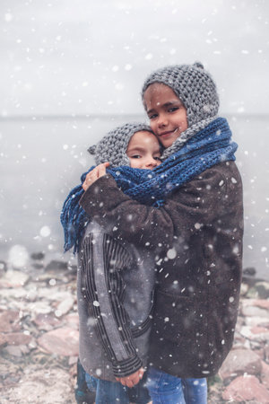 Little pretty girl in knitted grey hat hugging her frozen smaller brother during snowfall in winter, white cold weather, winter outside lifestyle close-up portraitの写真素材