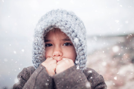 Little pretty girl in knitted grey hat during snowfall in winter, white cold weather, winter outside lifestyle close-up portraitの写真素材