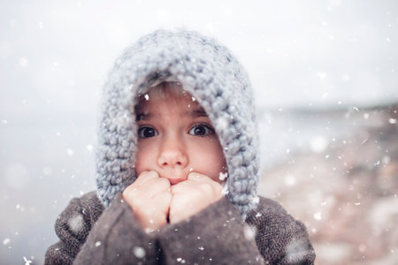 Little pretty girl in knitted grey hat during snowfall in winter, white cold weather, winter outside lifestyle close-up portraitの写真素材