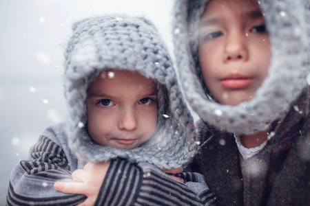 Little pretty girl in knitted grey hat hugging her frozen smaller brother during snowfall in winter, white cold weather, winter outside lifestyle close-up portraitの写真素材