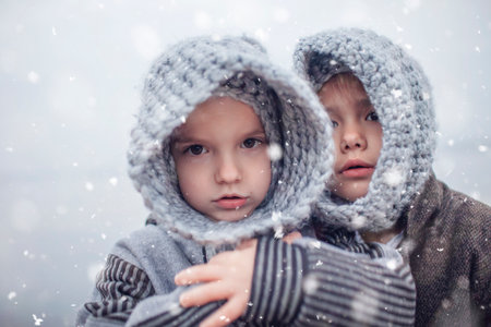 Little pretty girl in knitted grey hat hugging her frozen smaller brother during snowfall in winter, white cold weather, winter outside lifestyle close-up portraitの写真素材