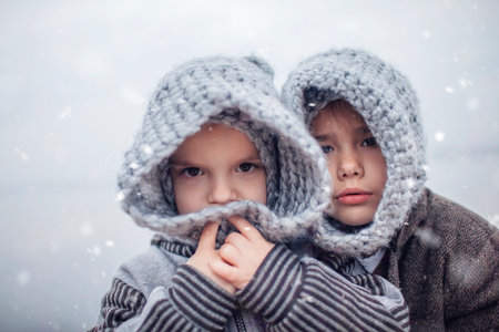 Little pretty girl in knitted grey hat hugging her frozen smaller brother during snowfall in winter, white cold weather, winter outside lifestyle close-up portraitの写真素材