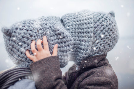 Little pretty girl in knitted grey hat hugging her frozen smaller brother during snowfall in winter, white cold weather, winter outside lifestyle close-up portraitの写真素材