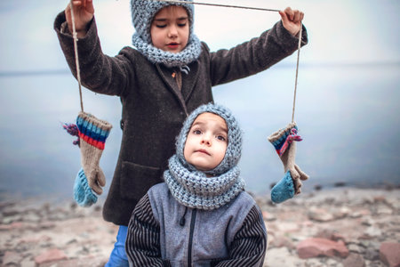 Little pretty girl in knitted grey hat proposes to share a pair of gloves with her frozen smaller brother during snowfall in winter, white cold weather, outside lifestyle portraitの写真素材