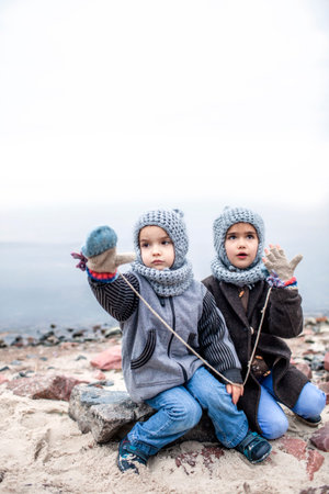 Little pretty girl in knitted grey hat sharing a pair of gloves with her frozen smaller brother during snowfall in winter, white cold weather, outside lifestyle portraitの写真素材