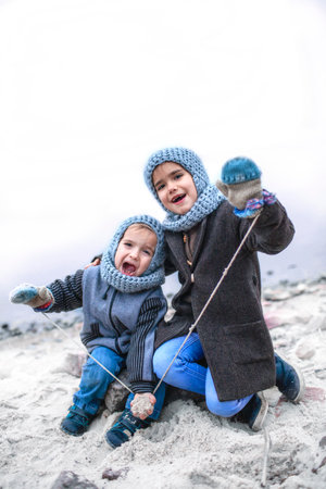 Little pretty girl in knitted grey hat sharing a pair of gloves with her frozen smaller brother during snowfall in winter, white cold weather, outside lifestyle portraitの写真素材