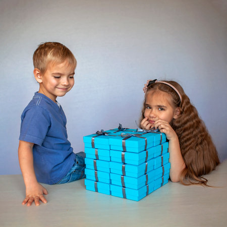 Cute little girl with her smaller brother near a huge pile of blue gift boxes with gray ribbons over white studio backgroundの写真素材