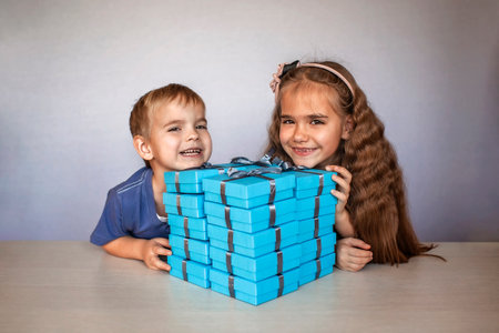 Cute little girl with her smaller brother near a huge pile of blue gift boxes with gray ribbons over white studio backgroundの写真素材
