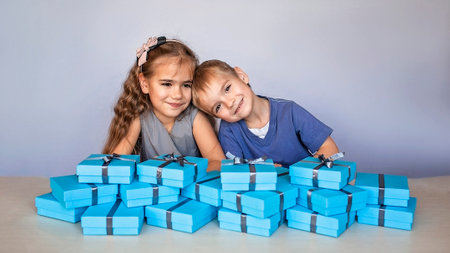 Cute little girl with her smaller brother near a huge pile of blue gift boxes with gray ribbons over white studio backgroundの写真素材